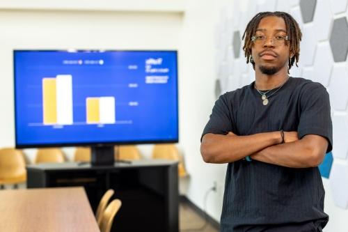 A student stands with their arms crossed in front a large computer monitor displaying a bar graph